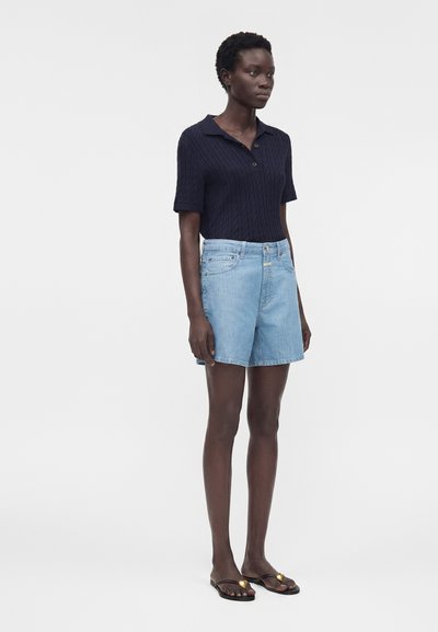 Γυναίκα με μπλε ναυτικό μπλουζάκι πόλο με κοντά μανίκια, ανοιχτό μπλε τζιν σορτς και μαύρες μπαλαρίνες με χρυσή λεπτομέρεια, στέκεται σε απλό φόντο.