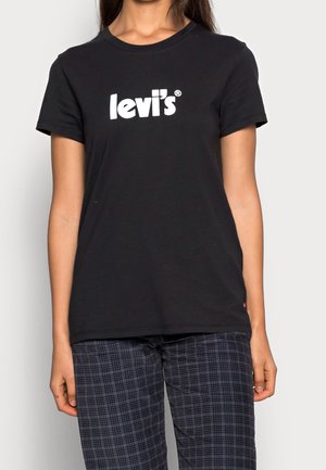 Femme portant un t-shirt noir Levi's avec un logo blanc et un pantalon à carreaux bleu foncé, debout devant un fond clair uni.