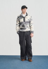 Νέος άνδρας που φοράει μαύρο καπέλο, πουκάμισο fleece με σχέδια, φαρδιά μαύρη παντελόνα cargo και καφέ δερμάτινα παπούτσια, στέκεται σε μπλε χαλί.