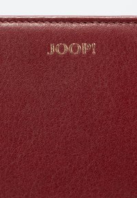 Cartera de cuero burdeos con superficie texturizada y logo "JOOP!" en relieve dorado. Bordes lisos, diseño compacto adecuado para el uso diario.