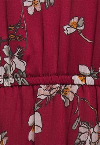 Tissu rouge avec un motif floral présentant des fleurs blanches et des centres jaunes, détail froncé à la taille, texture lisse.