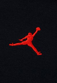 Silhouette rouge d’un joueur de basket sautant avec un bras levé tenant un ballon, brodée sur tissu noir.