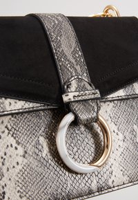 Černá semišová kabelka s texturovaným šedým hadím vzorem na klopě, která má kulatý zlatý a stříbrný kovový prvek.