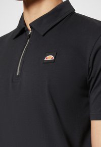 Gros plan d'une personne portant un polo noir à manches courtes avec une fermeture éclair argentée et un petit logo rouge et orange sur la poitrine.