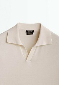 Chemise en maille beige clair avec un col doux et un décolleté en V, montrant une étiquette noire à l’intérieur du col.