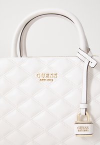 Bolso de mano acolchado blanco con un logo dorado y un candado decorativo. Fabricado en material sintético con patrones de diamante cosidos y asas suaves.
