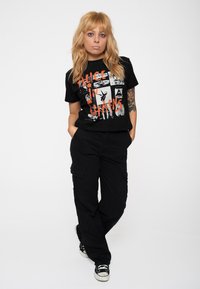 Black short-sleeve t-shirt featuring a graphic design with orange text and black and white images. Worn with black cargo pants and sneakers.