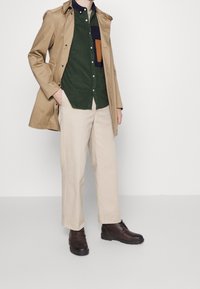 Beige trenchcoat, grön skjorta med knappar och färgblockeringsficka, ljusa byxor och mörkbruna stövlar. Enkelt mönster och tygtexturer.