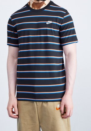 Mężczyzna w czarnym krótkim rękawie T-shircie Nike z niebiesko-pomarańczowymi poziomymi paskami oraz beżowych szortach Nike z pomarańczowym logo.