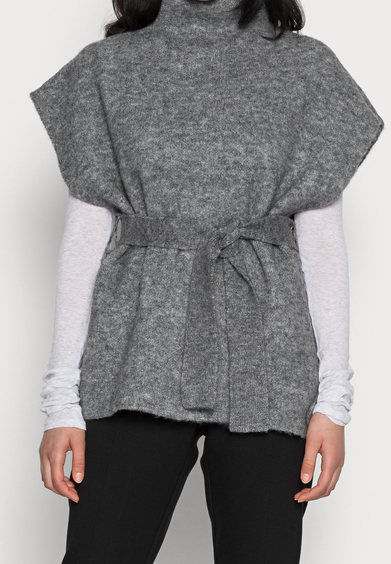 Femme portant un gilet sans manches en maille grise avec une ceinture nouée à la taille, par-dessus un haut blanc à manches longues et un pantalon noir.