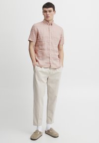 Lichtroze linnen overhemd met korte mouwen en een button-down kraag, gecombineerd met beige, iets loszittende broeken en beige klompen.