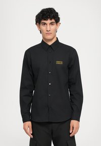 Svart skjorte med lange ermer, spiss krage, knappestolpe og gullmerke på brystet. Glatt stoff og tilpasset passform.