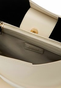 Borsetta in pelle crema con hardware della cerniera dorata, caratterizzata da una texture liscia e un'etichetta interna con la scritta "ENERGIA POSITIVA."