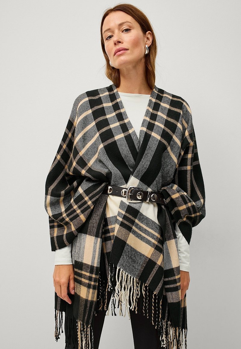 Oversized plaid poncho in black, beige, and gray with fringed edges, belted at the waist. Soft material and loose-fitting design.