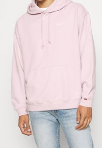 Sudadera rosa claro con un bolsillo canguro, capucha con cordón y pequeño logo blanco en el pecho. Textura suave, corte relajado y puños acanalados.