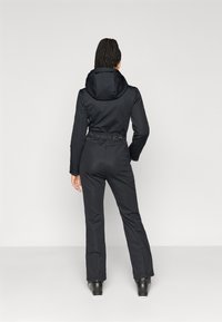 Μαύρη φόρμα με εφαρμοστό σχέδιο, κουκούλα και ζωνάκι στη μέση. Διαθέτει μακριά μανίκια και φερμουάρ στα μπατζάκια. Λείος υφή και μοντέρνα σιλουέτα.