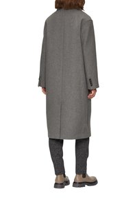 Lange grijze wollen jas met een rechthoekige snit, verlaagde schouders en knoopmanchetten, gecombineerd met gestructureerde antracietkleurige broek en bruine laarzen.