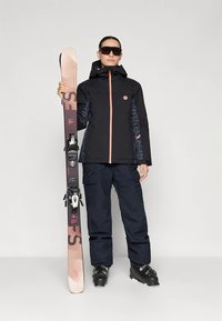 Czarna i wzorzysta kurtka narciarska z pomarańczowymi akcentami, granatowe spodnie, trzyma narty i nosi okulary przeciwsłoneczne, prezentując zimowy strój sportowy.