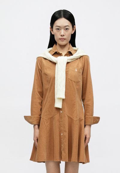 Καφέ βαμβακερό φόρεμα με μακριά μανίκια, κουμπιά μπροστά και πλισέ φούστα· συνδυασμένο με λευκό πουλόβερ με πλεκτό σχέδιο καλωδίου, δεμένο στο λαιμό.