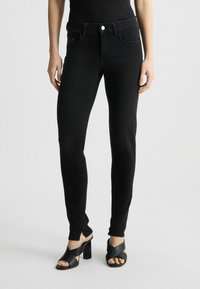 Crne traperice skinny s srednjim strukom, prednjim džepovima i suptilnom teksturom. Uparene s crnim sandalama na otvorenom prstiju koje imaju blok potpeticu.