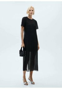 Črna plisirana maksi obleka s kratkimi rokavi, prozoren material, v kombinaciji z majhno črno torbico z belimi žebljički in srebrnimi sandali s peto.