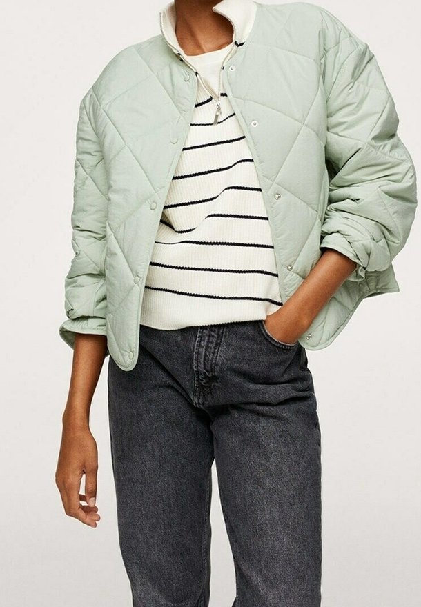 Personne portant une veste matelassée vert clair par-dessus un pull rayé blanc et noir, avec un jean gris foncé taille haute, main dans la poche.