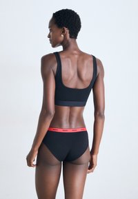 Femme vue de dos portant un soutien-gorge de sport noir et un sous-vêtement noir avec une taille rouge étiquetée "HUGO" sur un fond blanc uni.