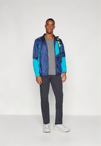 Muška jakna u mornarsko plavoj i tirkiznoj boji, s prednjim patentnim zatvaračem i kapuljačom, u kombinaciji sa sivom majicom i tamnim hlačama. Obuća su svijetle tenisice.