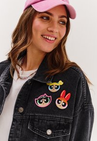 Czarna dżinsowa kurtka z trzema kolorowymi haftowanymi naszywkami z postaciami z kreskówek po lewej stronie. Włożona różowa czapka z daszkiem.