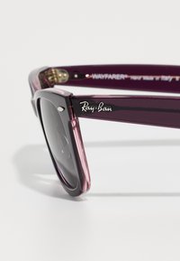 Ljubičaste prozirne Ray-Ban Wayfarer naočale s tamnim lećama prikazane izbliza na bijeloj površini.