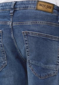 Primer plano de la parte trasera de unos vaqueros vaqueros azules con costuras visibles, bolsillos traseros, trabillas para cinturón y una etiqueta de cuero marrón con la etiqueta "INDICODE EST. 1993."