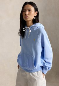 Jeune femme portant un sweat à capuche bleu clair avec des cordons blancs et un pantalon beige, debout devant un fond beige neutre.