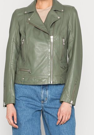 Veste de biker en cuir vert olive avec fermeture éclair asymétrique à l'avant, poches zippées et accessoires argentés, portée sur un top beige et un jean bleu.