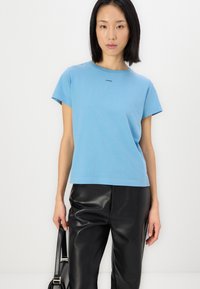 Ανοιχτό μπλε βαμβακερό μπλουζάκι με κοντά μανίκια και λαιμόκοψη, με ένα μαύρο λογότυπο στο μπροστινό μέρος. Συνδυασμένο με μαύρο δερμάτινο παντελόνι.