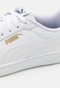 Baskets en cuir blanc avec détails texturés, lacets plats blancs, logo "PUMA" doré et semelle en caoutchouc avec marquage en relief.