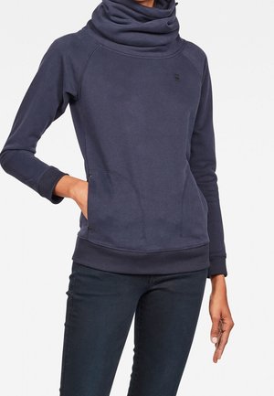 Femme portant un pull à manches longues bleu foncé avec un grand col bénitier et des poches à l’avant, associé à un pantalon foncé.