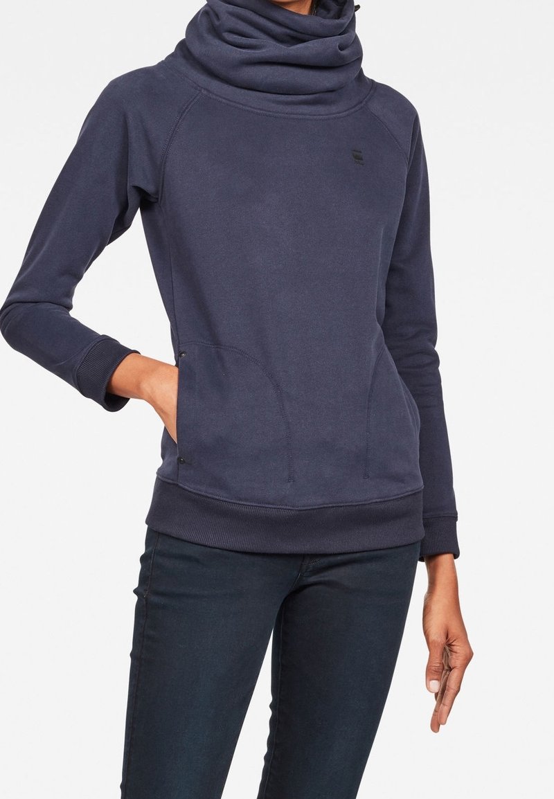 Femme portant un pull à manches longues bleu foncé avec un grand col bénitier et des poches à l’avant, associé à un pantalon foncé.