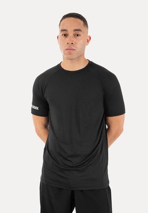 Mann mit kurzen Haaren und Sommersprossen, der ein schlichtes schwarzes kurzärmeliges Shirt und schwarze Hose trägt, steht mit den Händen hinter dem Rücken vor weißem Hintergrund.