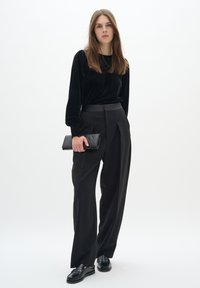Črna žametna bluzica z dolgimi rokavi v kombinaciji z visokim pasom črnimi hlačami. Model drži črn nahrbtnik ter nosi črne loafers čevlje. Minimalistična zasnova.