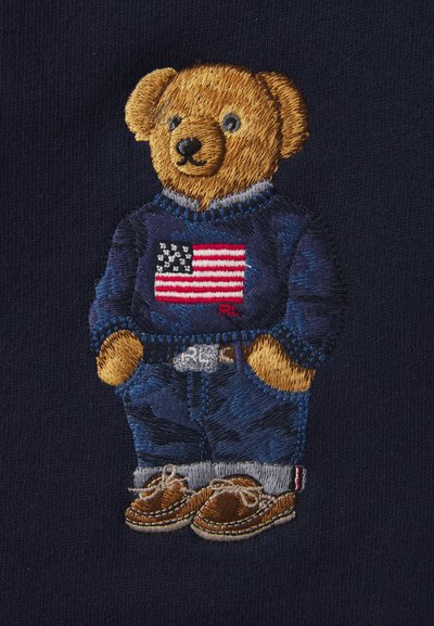 Panda en peluche brodé portant un pull marine avec un motif de drapeau américain, un jean bleu et des chaussures brunes avec lacets. Traits du visage détaillés.