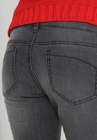 Tummanharmaat farkut, joissa on vartalonmyötäinen muotoilu, kahdella takataskulla ja näkyvillä ompeluyksityiskohdilla. Kangas on hieman pintakuvioitu.