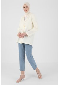Cream-colored blazer with two-button closure, relaxed fit. Paired with light blue jeans and strappy beige heels. Smooth fabric texture.
