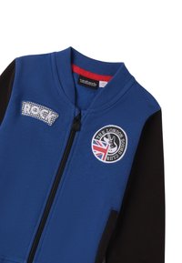 Chaqueta azul con cremallera y mangas negras. Presenta parches bordados: "ROCK" y "THE LONDON SOUND CLUB" con un diseño de guitarra y la bandera del Reino Unido.