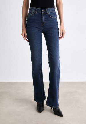 Osoba nosząca ciemnoniebieskie, dżinsy z wysokim stanem i prostą nogawką, połączone z czarnymi butami na obcasie z ostrym noskiem na beżowej podłodze.