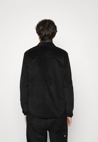 Camicia in velluto a coste nera con scollo a colletto, maniche lunghe e orlo dritto, caratterizzata da una texture sottile e senza motivi o accenti visibili.