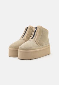 Stivaletti beige in suede a caviglia con chiusura a zip, dotati di una suola piatta spessa e dettagli cuciti per un tocco extra di texture.
