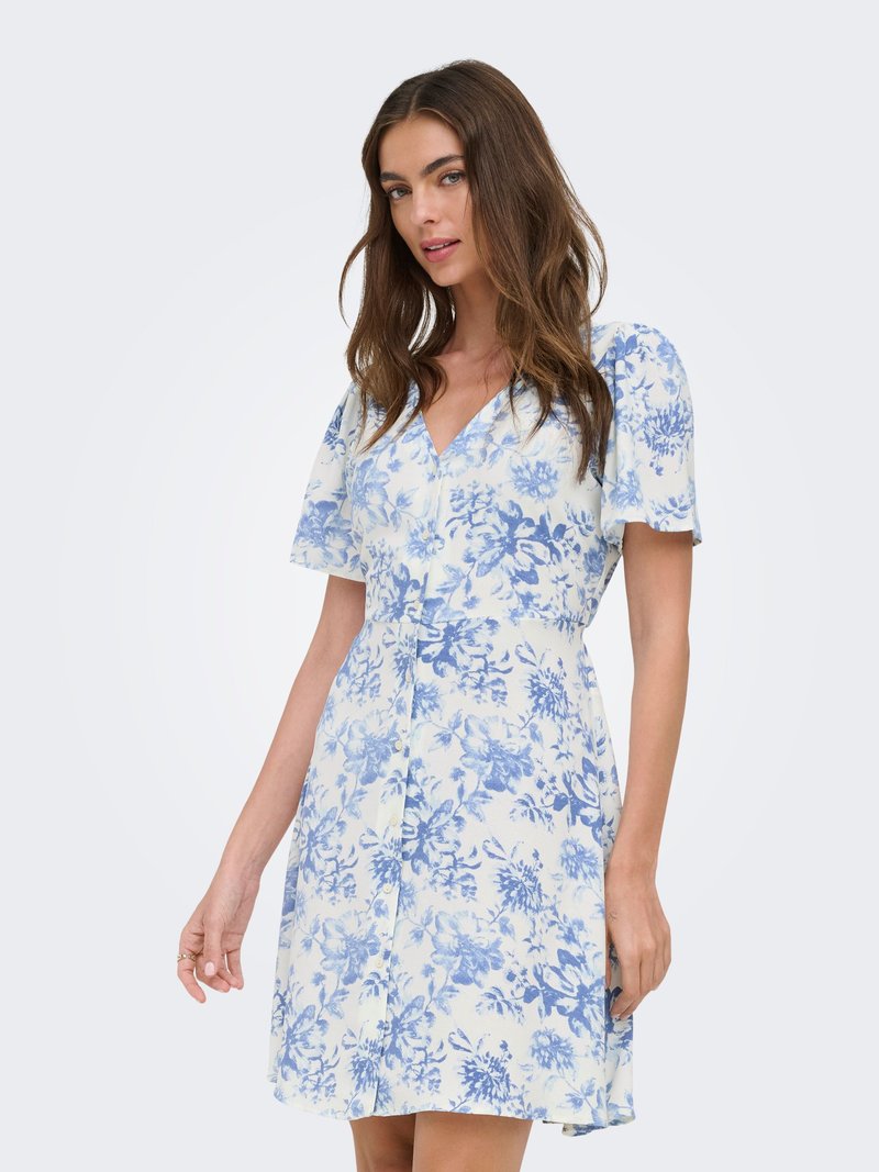 Vrouw die een witte jurk draagt met een blauwe bloemenprint, korte mouwen en knopen aan de voorkant, staand tegen een effen achtergrond.