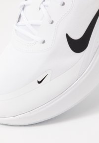 Vit sportskor med en slät yta, svart Nike-swoosh och vita snören. Har en rund tå och en hållbar sula.