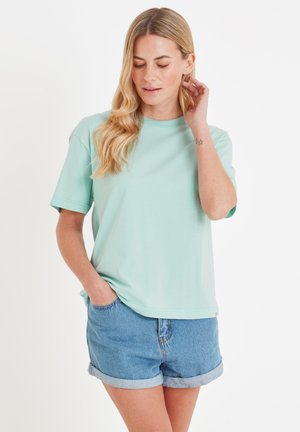 Woman with long blonde hair wearing a mint green t-shirt and rolled-up denim shorts, with one hand in pocket and the other touching her ear.