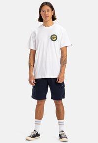 Camiseta de algodón blanca con un parche de logo circular en amarillo y negro en el pecho, combinada con pantalones cortos cargo azul marino y zapatillas negras.